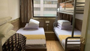 A compact hotel room with two beds, one with a checkered bedspread and the other with a plain bedspread. There is a window with curtains, a small bedside table, and a wall-mounted light. - Lucky Guest House (Kowloon)