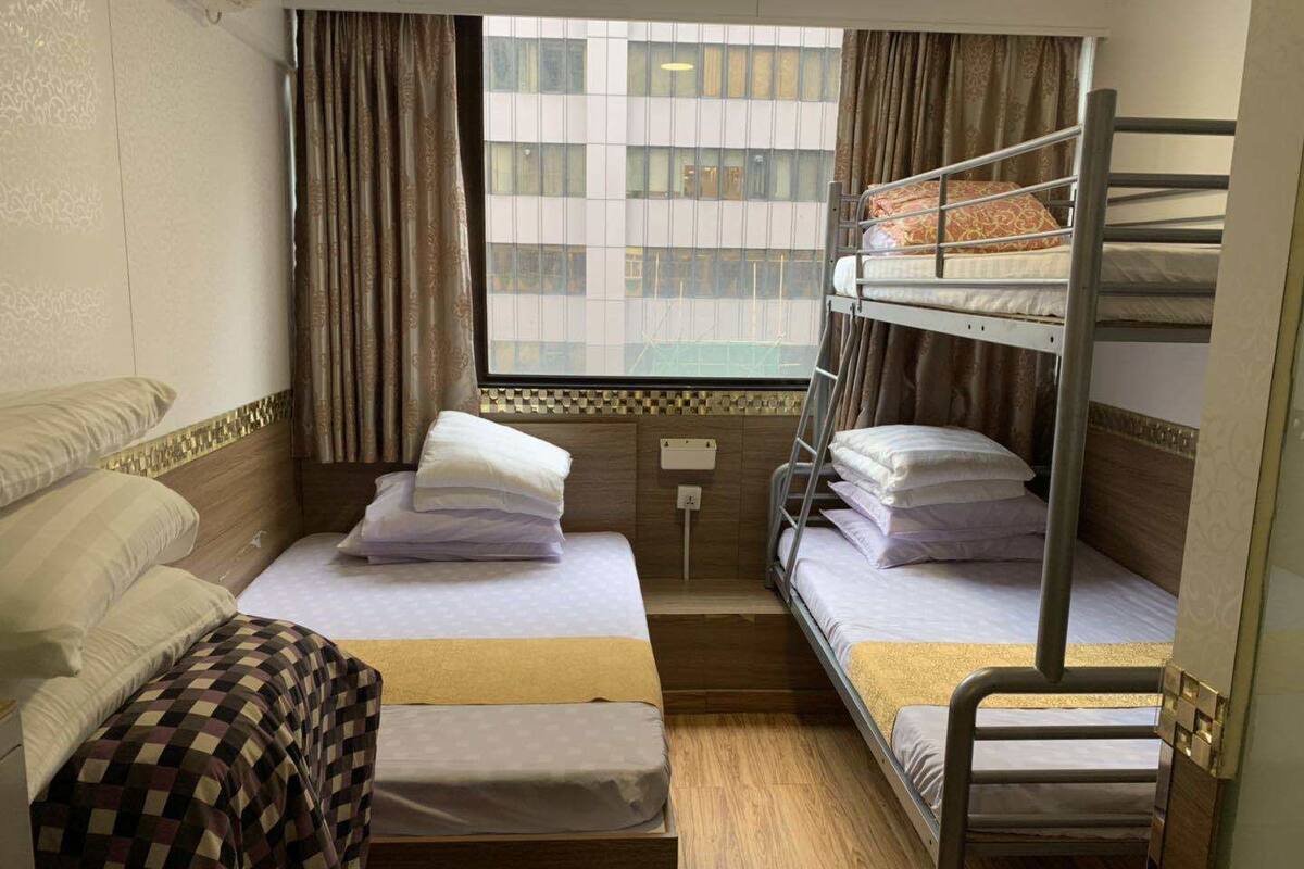 A compact hotel room with two beds, one with a checkered bedspread and the other with a plain bedspread. There is a window with curtains, a small bedside table, and a wall-mounted light.