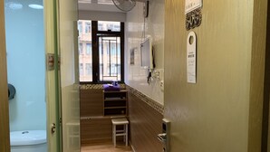 A hotel room with a wooden floor, a window with a view of buildings, a small stool, a shelf with towels, and a door with a security lock. - Lucky Guest House (Kowloon)