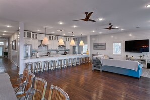 A modern kitchen with white cabinets, a central island with bar stools, a dining area with a blue sofa, and a TV mounted on the wall.