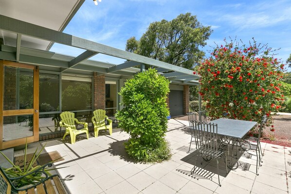 A covered patio area with a table and chairs, a small tree, and a bush. - Charlton House (Aireys Inlet)