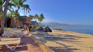Ein Hotel am Strand mit Liegestühlen, Palmen und Blick auf das Meer und die Berge.