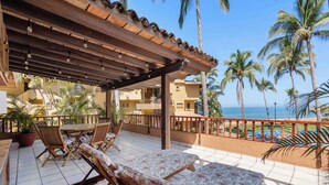 Ein überdachter Sitzbereich im Freien mit Lounge-Stühlen und einem Tisch, mit Blick auf einen Strand mit Palmen und eine klare Meerblick.