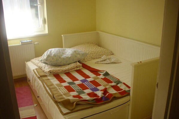 Una habitación pequeña con una cama individual, cabecera blanca sencilla, una ventana con cortina y un radiador debajo.