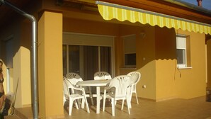 Een terras buiten met een ronde tafel en witte stoelen, een geel zonnescherm en een raam met luiken.