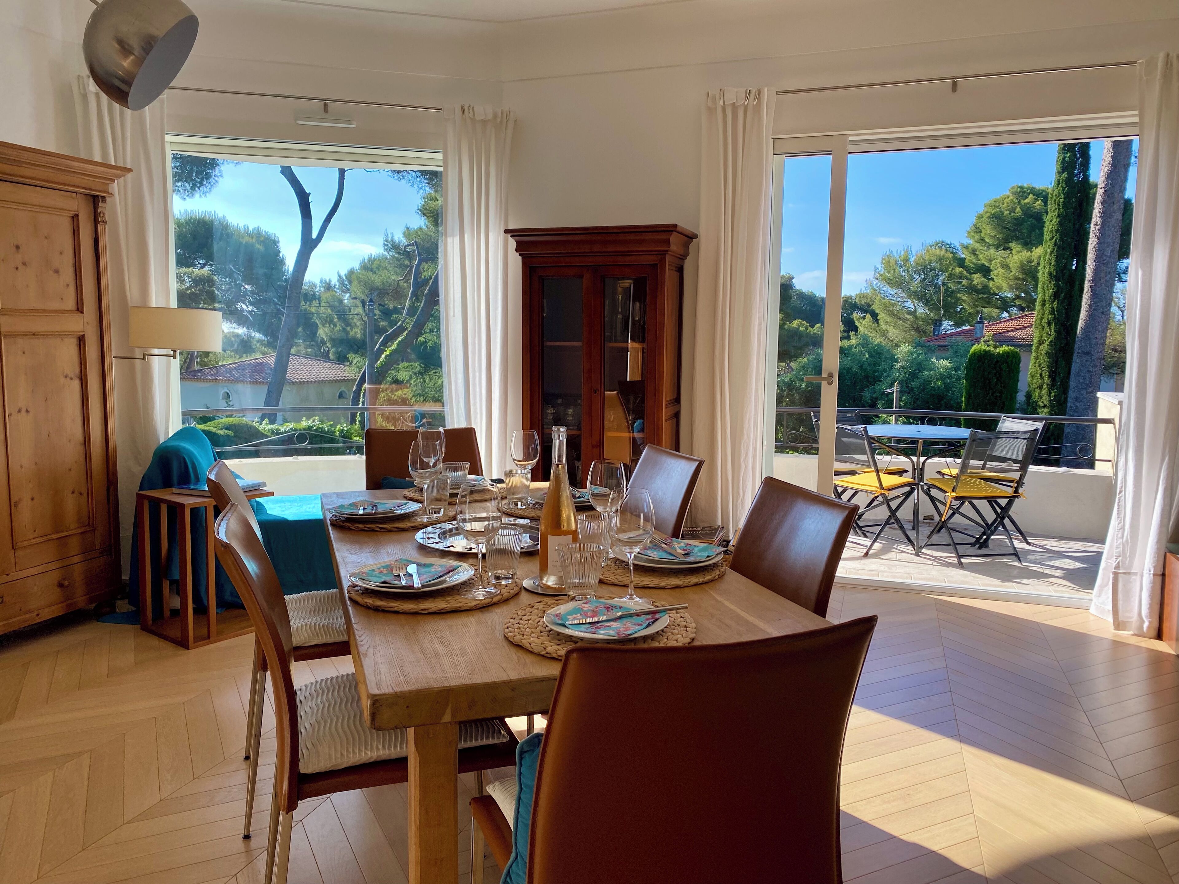 A dining area with a wooden table set for four, surrounded by chairs. There is a view of the outdoors through large windows.