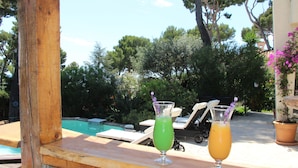 Dos bebidas sobre una mesa de madera junto a una piscina con sillones de descanso y una planta en maceta al fondo.