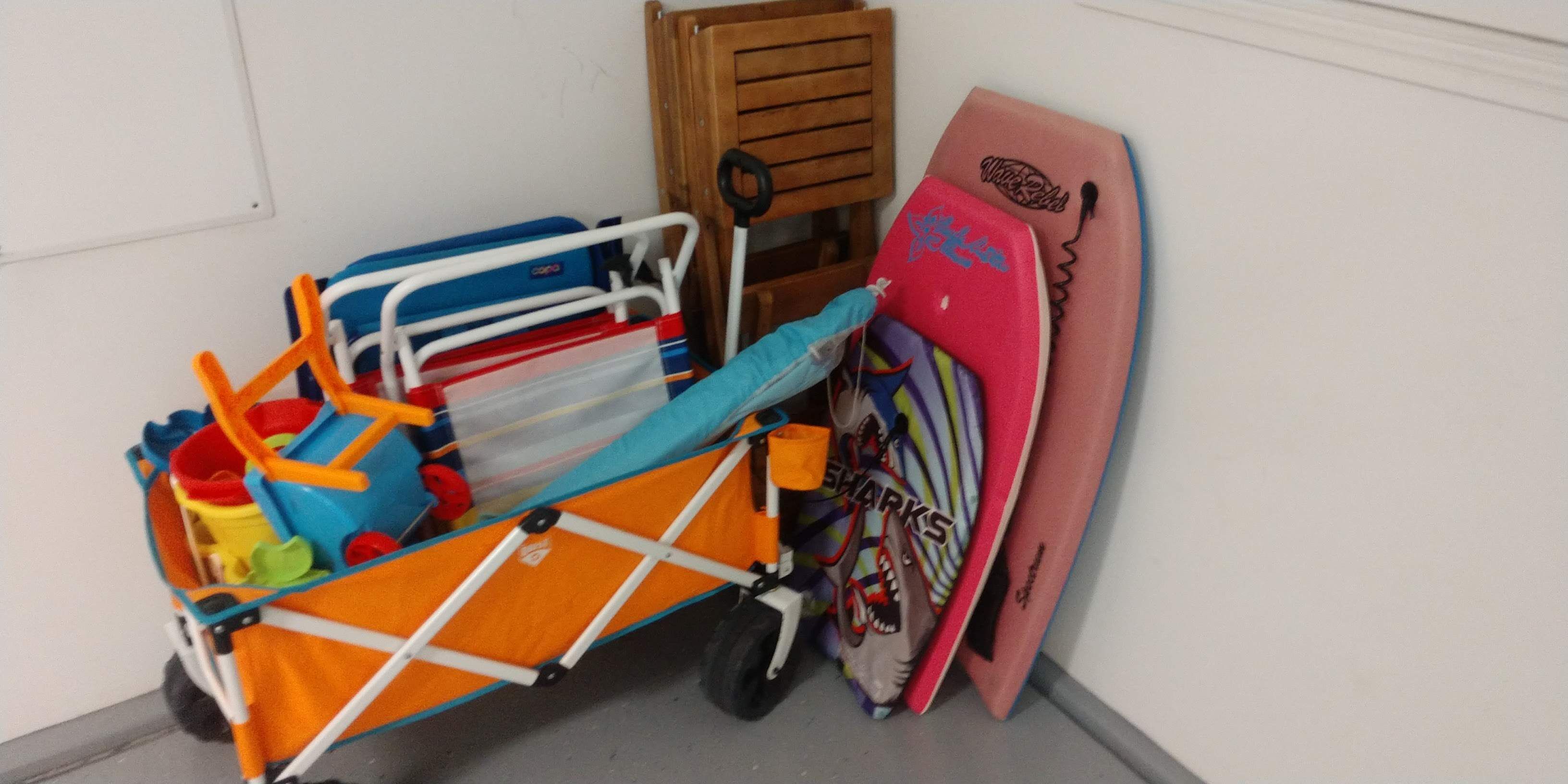 Un chariot contenant divers objets, notamment une serviette bleue, un seau rouge et une planche de surf rose, appuyée contre un mur.