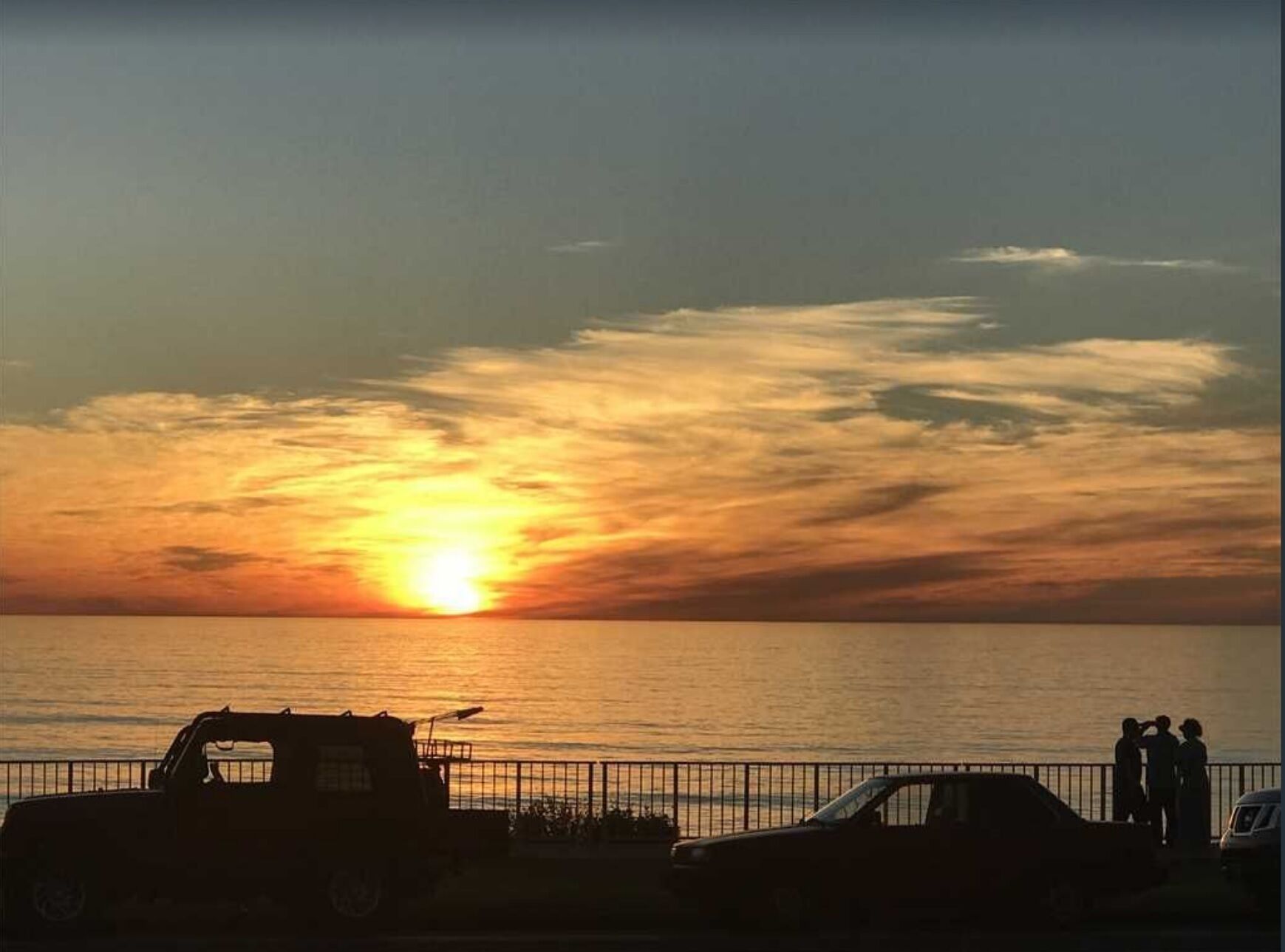 Ein Sonnenuntergang über dem Meer mit Silhouetten von Fahrzeugen und Menschen am Strand.