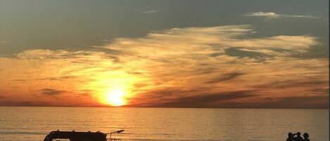 Ein Sonnenuntergang über dem Meer mit Silhouetten von Fahrzeugen und Menschen am Strand.