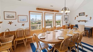A dining area with a round wooden table, multiple chairs, and a view through large windows. - Semi-Oceanfront Dog Friendly Home in Avon w/ Pool, Hot Tub, Tiki Bar, Game Room (Avon)