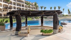 Een zwembadterras met een houten pergola, planten in potten en een uitzicht op een hotel in de verte.