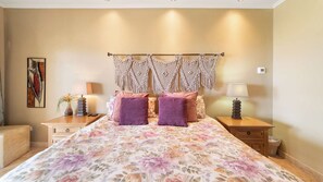 A bedroom with a floral bedspread, two purple pillows, a macrame wall hanging, bedside lamps, a small plant, and a framed artwork on the wall.