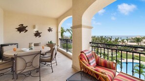 A balcony with a dining table and chairs, a wicker sofa, and a view of a pool and palm trees.
