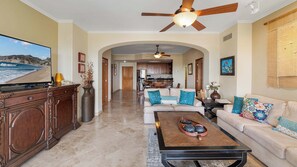 A spacious living room with a large flat-screen TV, a ceiling fan, and a kitchen in the background.