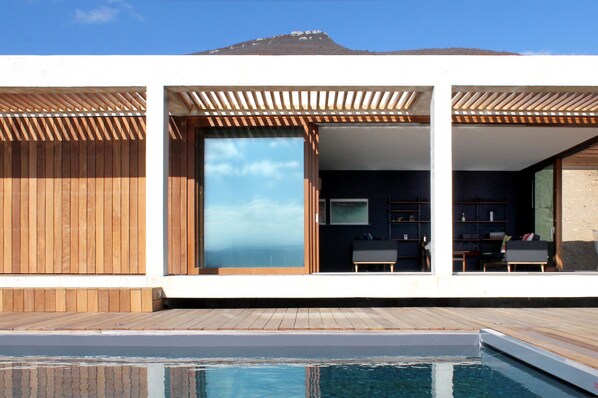 A modern outdoor pool area with wooden decking, a clear sky, and a mountain in the background.