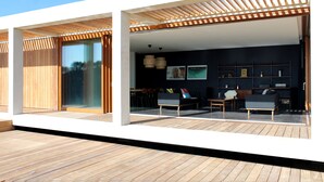 A modern outdoor living area with wooden flooring, a covered seating area with sofas and tables, and a dark wall with framed pictures.