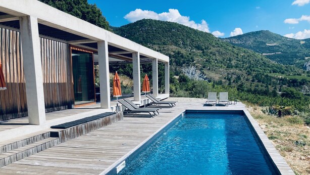 A modern outdoor pool with wooden decking, lounge chairs, and umbrellas, surrounded by lush greenery and mountains.
