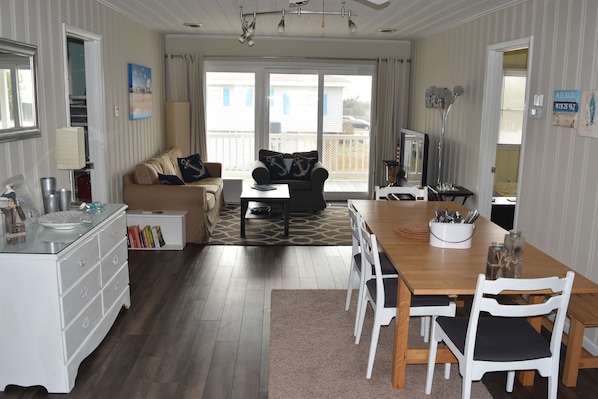 A well-lit living room with a sofa, dining table, and chairs. There is a kitchen area with a stove and cabinets. The room has large windows and a view of the outside.