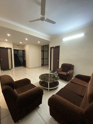 A spacious living area with a ceiling fan, a round coffee table with a vase of flowers, and brown armchairs. - Krishna Kunja Guest House (Bidhannagar)