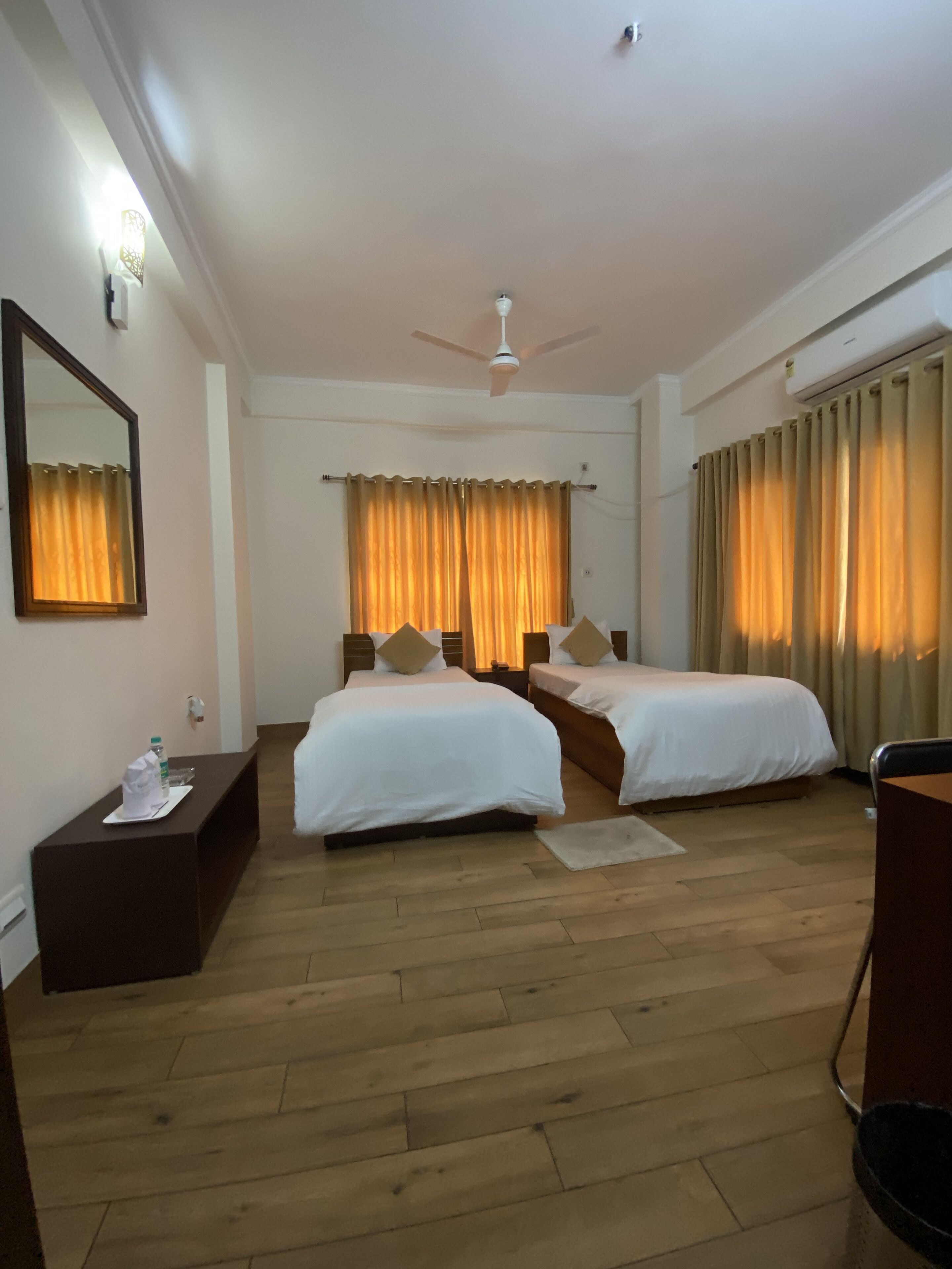 A hotel room with two beds, a ceiling fan, a desk with a chair, and a large mirror on the wall.