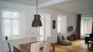 A modern living room with a wooden dining table, beige chairs, a black pendant light, a grey sofa, a TV on a wooden stand, and a window with curtains.