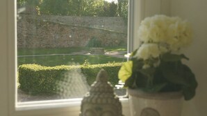 A window with a view of a stone wall and a fountain, a Buddha statue, and a potted plant with flowers.