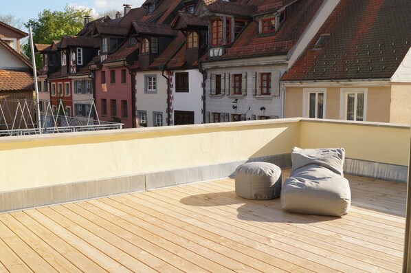 Terraza en la azotea con suelo de madera, dos sillones informales grises y vistas a edificios europeos tradicionales.