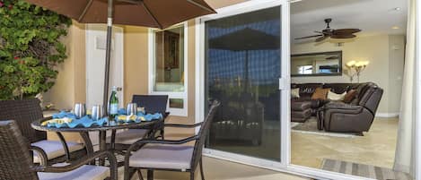 Un patio al aire libre con mobiliario de mimbre, una mesa con vasos y una botella, y un ventilador de techo visible a través de un ventanal.