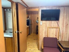 A narrow hallway with wooden walls, a sink, a refrigerator, a television mounted on the wall, and a small table with a chair.