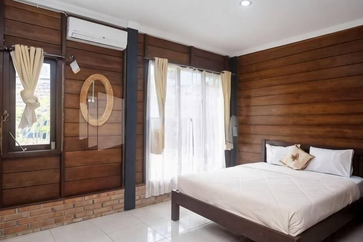 Een slaapkamer met een houten wand, een bed, een raam met gordijnen en een ronde spiegel.