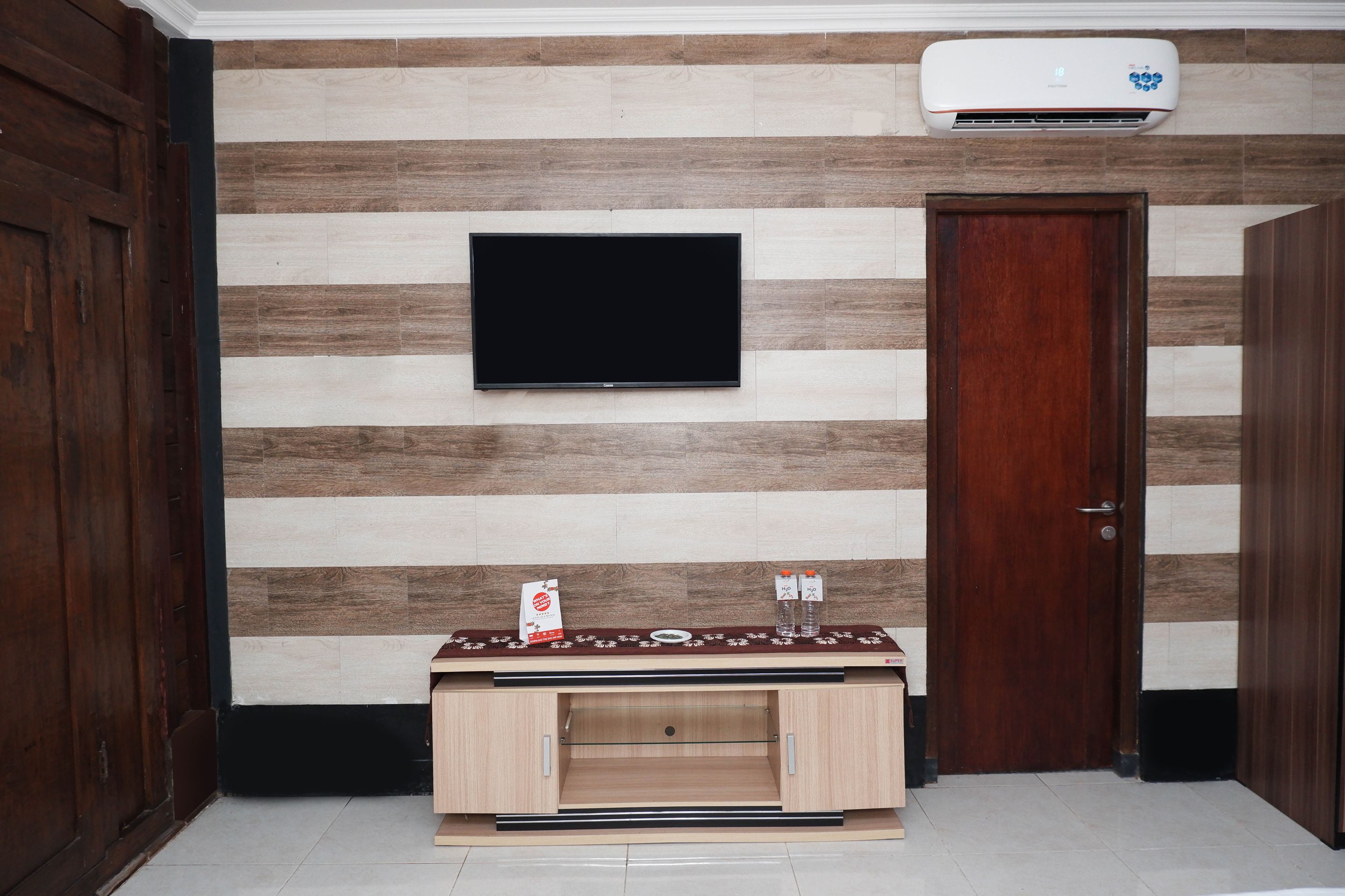A room with a flat-screen TV mounted on a wall with wooden paneling, a wooden door, and a small wooden cabinet with a decorative top.