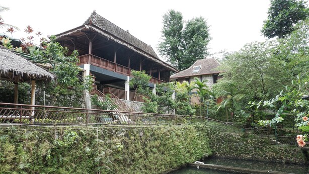 Un bâtiment traditionnel en bois, doté d’un toit de chaume, entouré d’une végétation luxuriante et traversé par un petit canal.