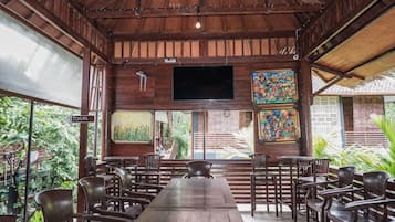 Interior de madera con una larga mesa y sillas, un televisor montado en la pared y un letrero que indica la ubicación de los baños.
