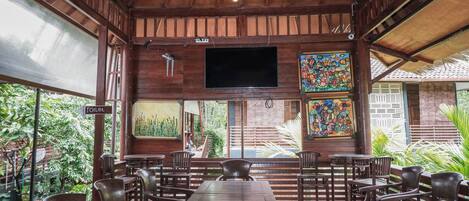 Interior de madera con una larga mesa y sillas, un televisor montado en la pared y un letrero que indica la ubicación de los baños.