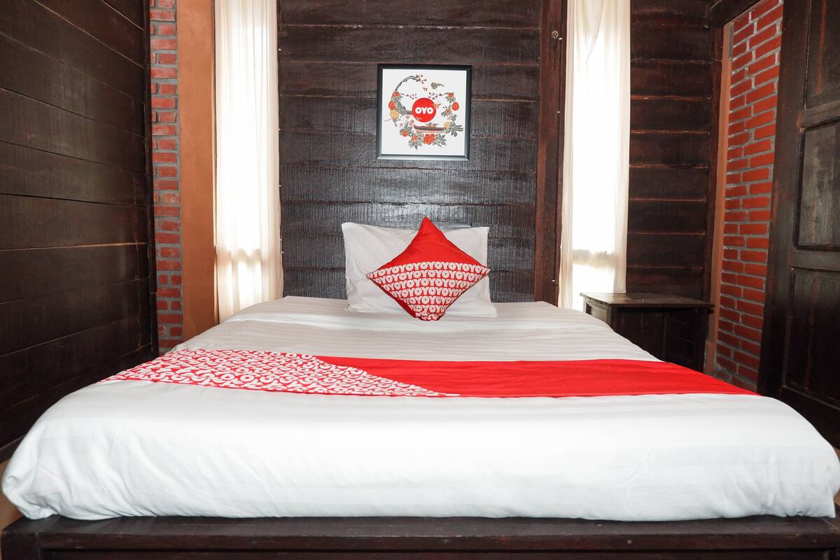 Een netjes opgemaakt bed met een wit-rode sprei, een kussen met een rood geometrisch patroon, en een lijst met een kunstwerk aan de muur erboven.