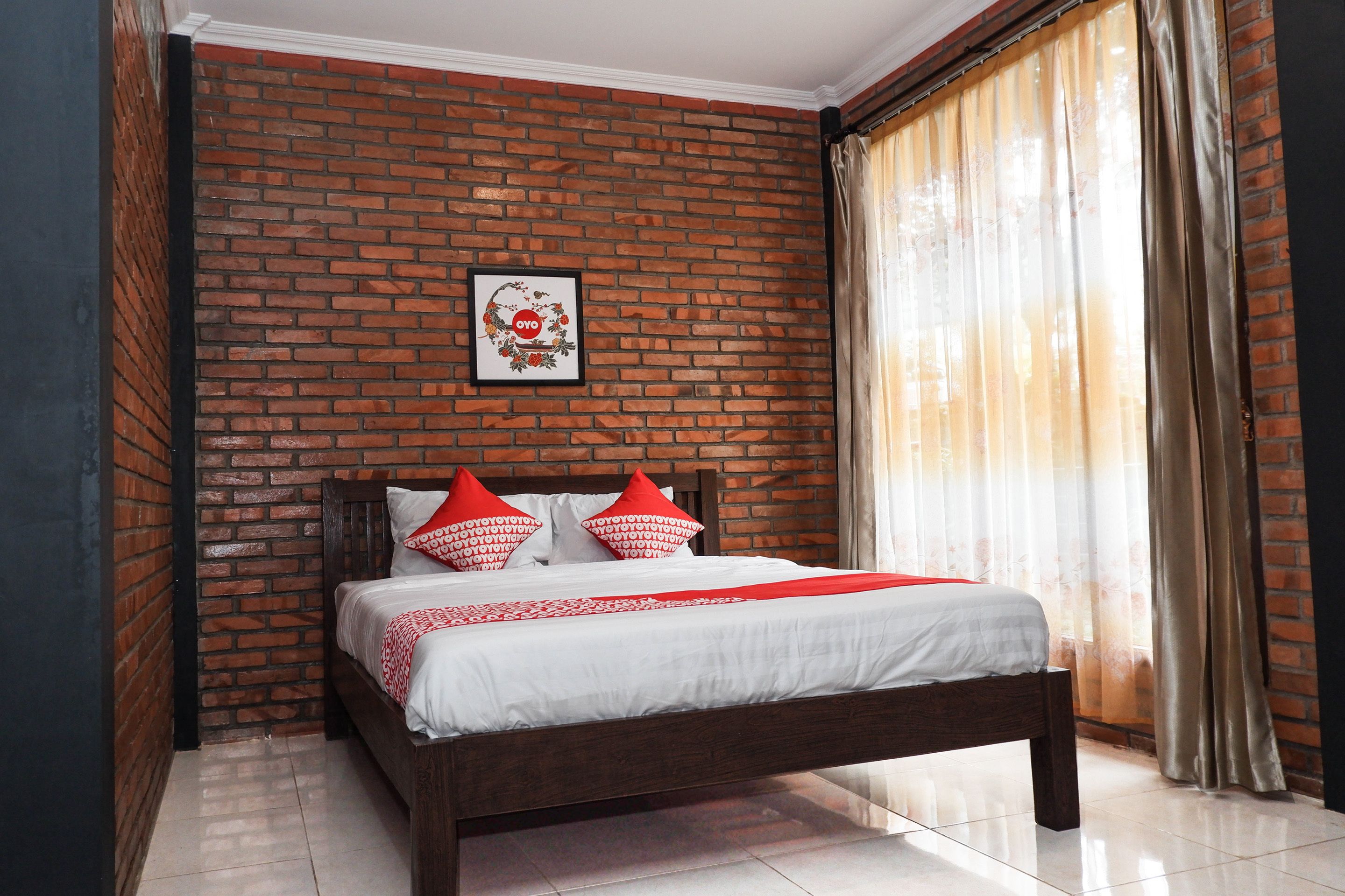 A bedroom with a brick wall, a bed with white bedding and red pillows, a framed picture on the wall, and a window with curtains.