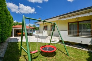 Ein Garten mit Schaukel, einer Steinterrasse und einem Haus mit rotem Dach und gelben Wänden.