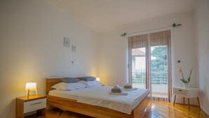 A neatly made bed with pillows and rolled towels, a bedside table with a lamp, a small white nightstand, a white furry rug, a window with curtains, and a small table with a vase of flowers. - Villa Tea in Imotski, private pool (Zmijavci)