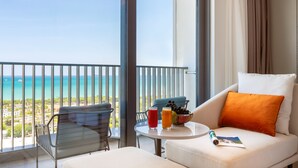 Een balkon met uitzicht op zee, een witte bank met een oranje kussen, een glazen tafel met een drankje en fruit, en een stoel.