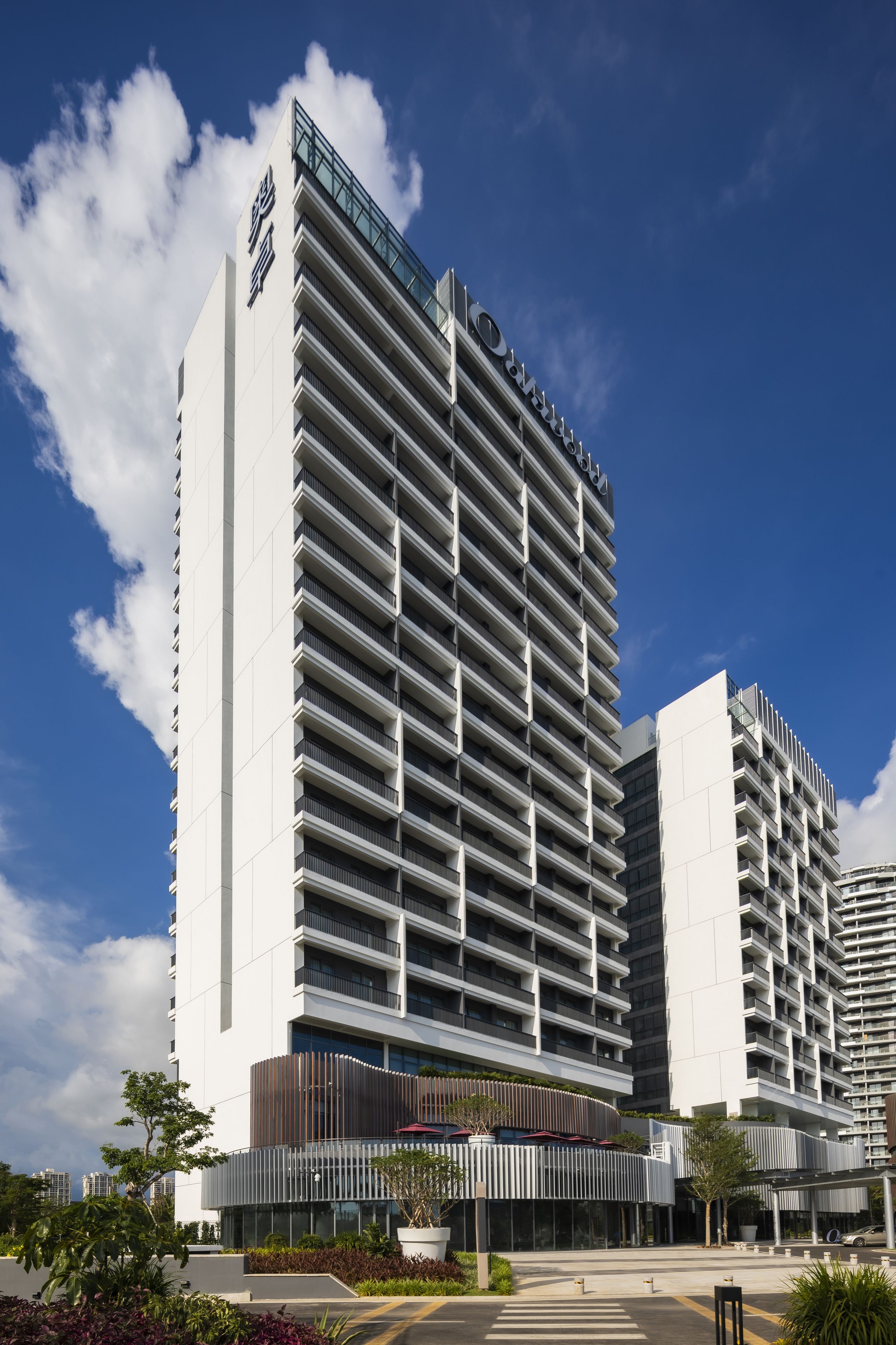 Un hotel moderno con un llamativo letrero vertical, varios balcones y un área de entrada ajardinada.