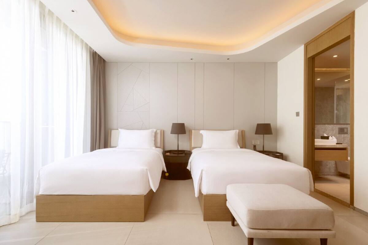 Une chambre d’hôtel avec deux lits, un banc, une table de chevet, une lampe et une salle de bain visible à travers une porte ouverte.