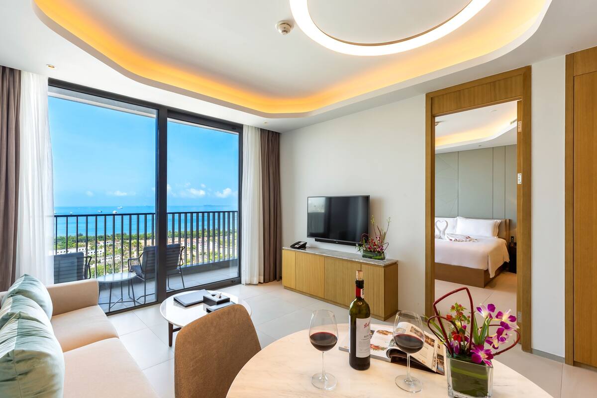 Une chambre d’hôtel moderne dotée d’un balcon, d’une télévision, d’un lit et d’un coin repas agrémenté d’une bouteille de vin et de fleurs.