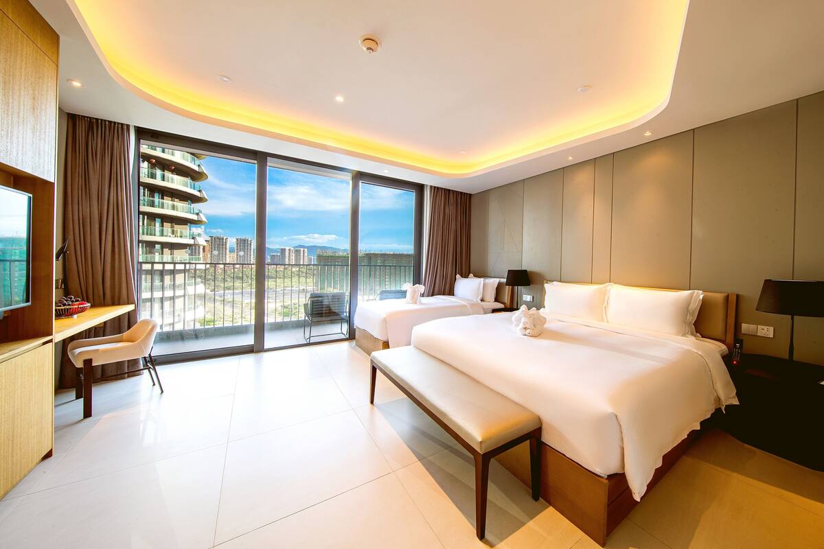 Une chambre d’hôtel moderne avec deux lits, un bureau et un grand balcon offrant une vue sur la ville.