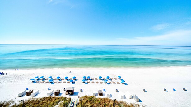 Een strand met wit zand en helderblauw water. Er staan rijen blauwe parasols en ligstoelen.