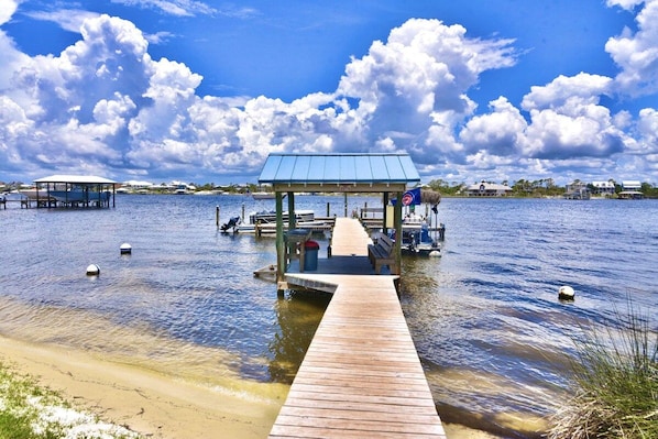 A wooden pier leading to a covered area with benches and a flag, surrounded by calm water and distant buildings.
