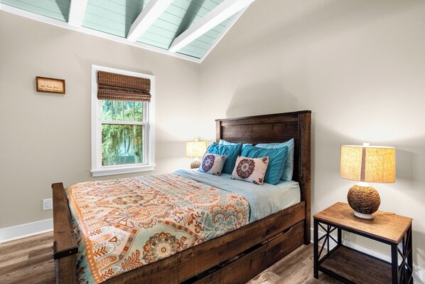 Un dormitorio con cama de madera, ropa de cama estampada, lámpara de noche, ventana con persianas y tragaluz.