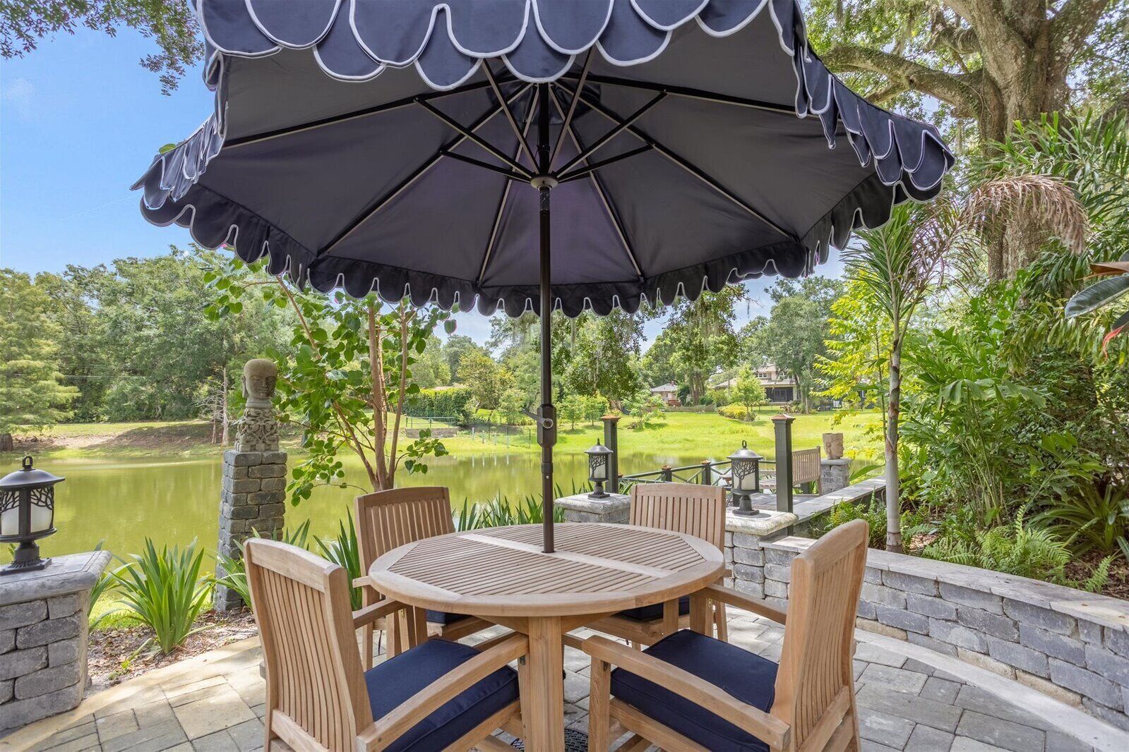 A patio with a round table and chairs, an umbrella, and a view of a lake and greenery.