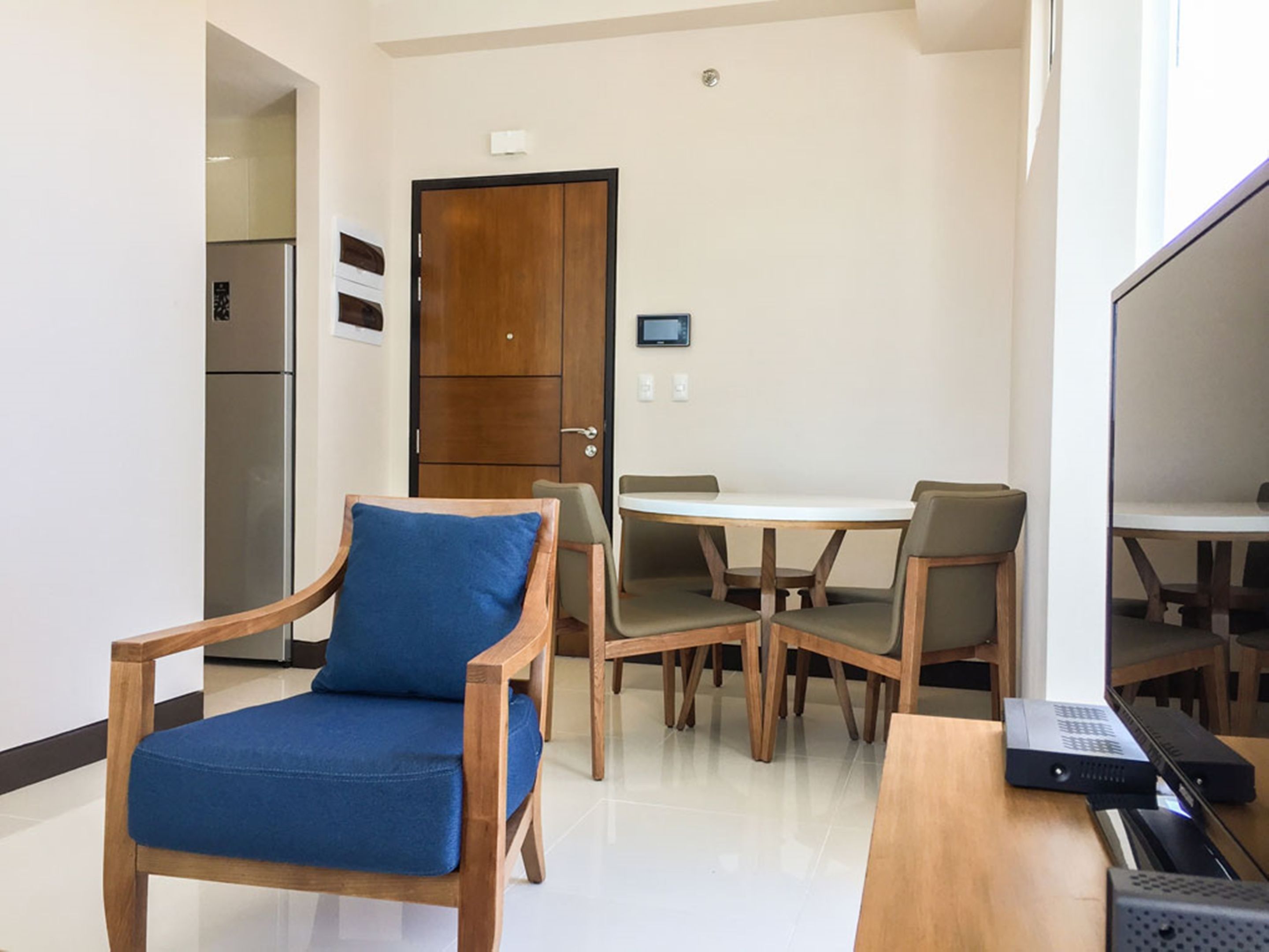 A modern hotel room with a wooden chair with a blue cushion, a dining table with four chairs, a refrigerator, and a flat-screen TV.