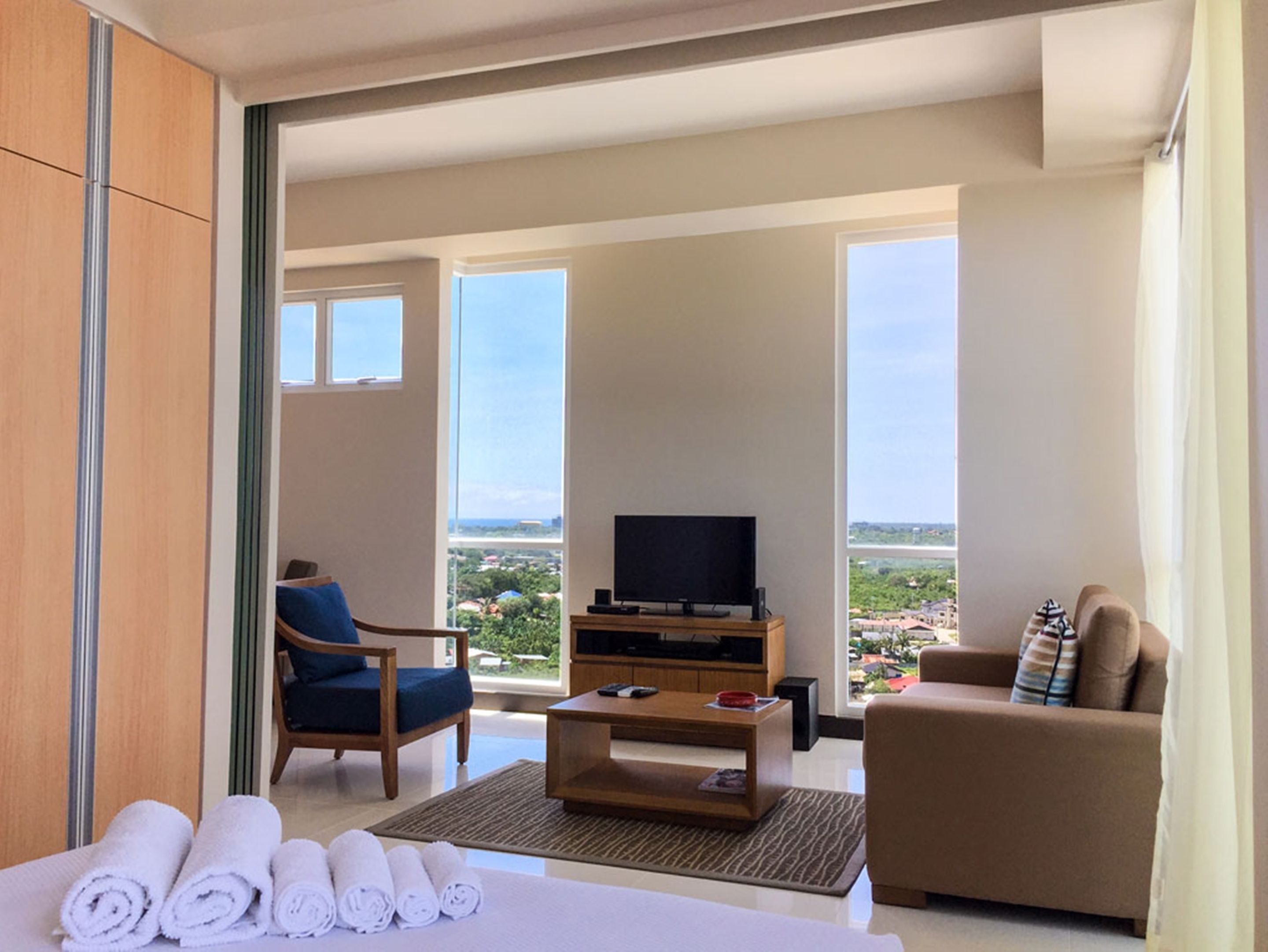 Quarto de hotel com varanda, televisão, sofá, cadeira e toalhas sobre uma mesa.